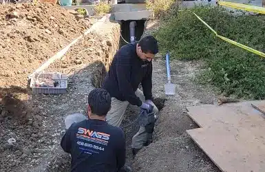Swags-Technicians-At-Work (1) Vertical view of two plumbers installing piping in a deep trench by a backyard deck