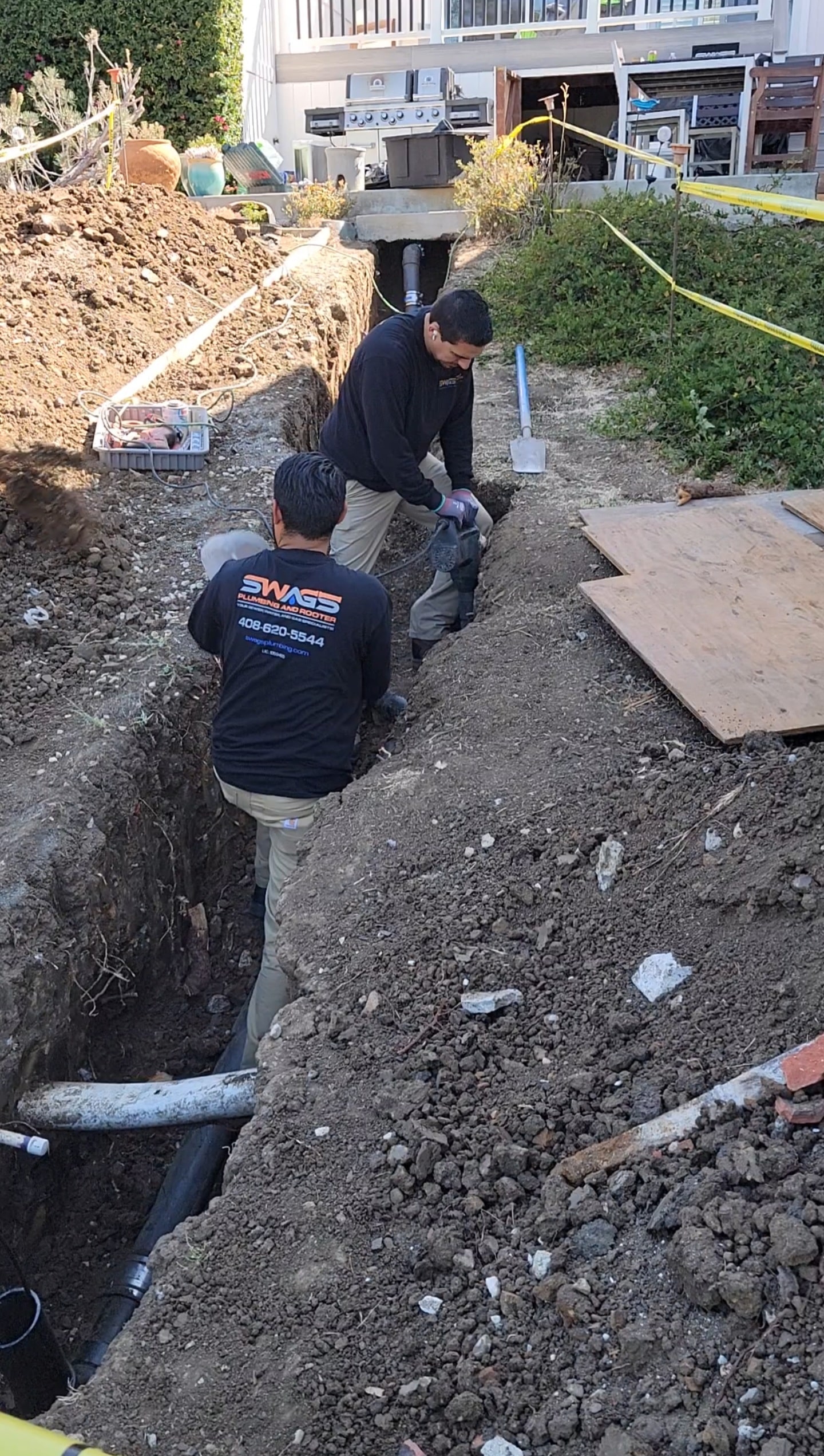 Swags Technicians At Work Two plumbers working in a dirt trench near a foundation with visible drainage pipe